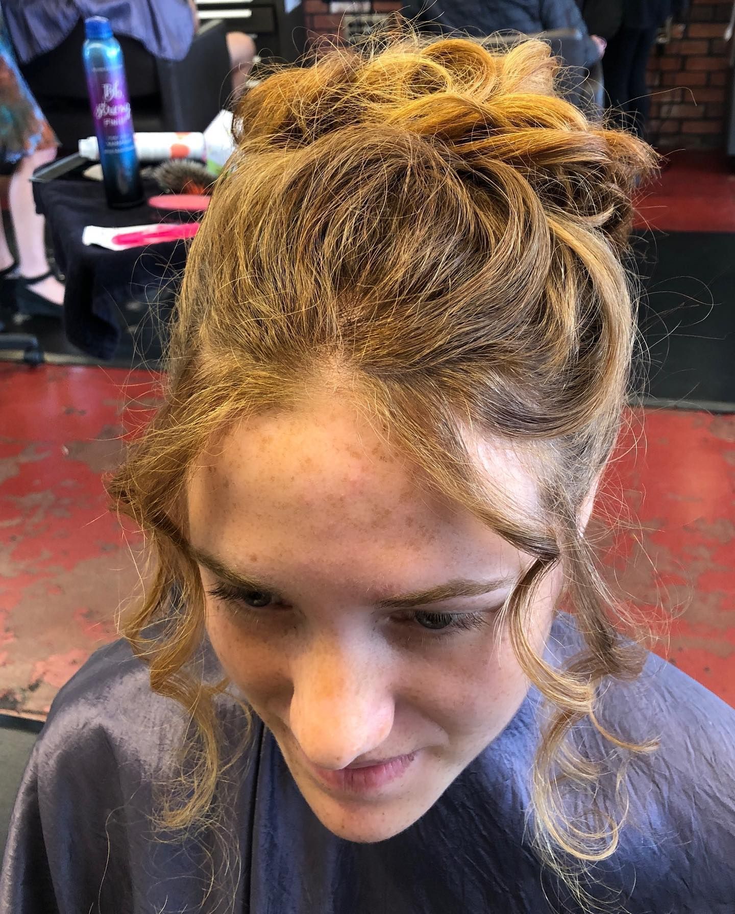 Person with light brown curly hair in an updo, wearing a blue cape, in a salon.