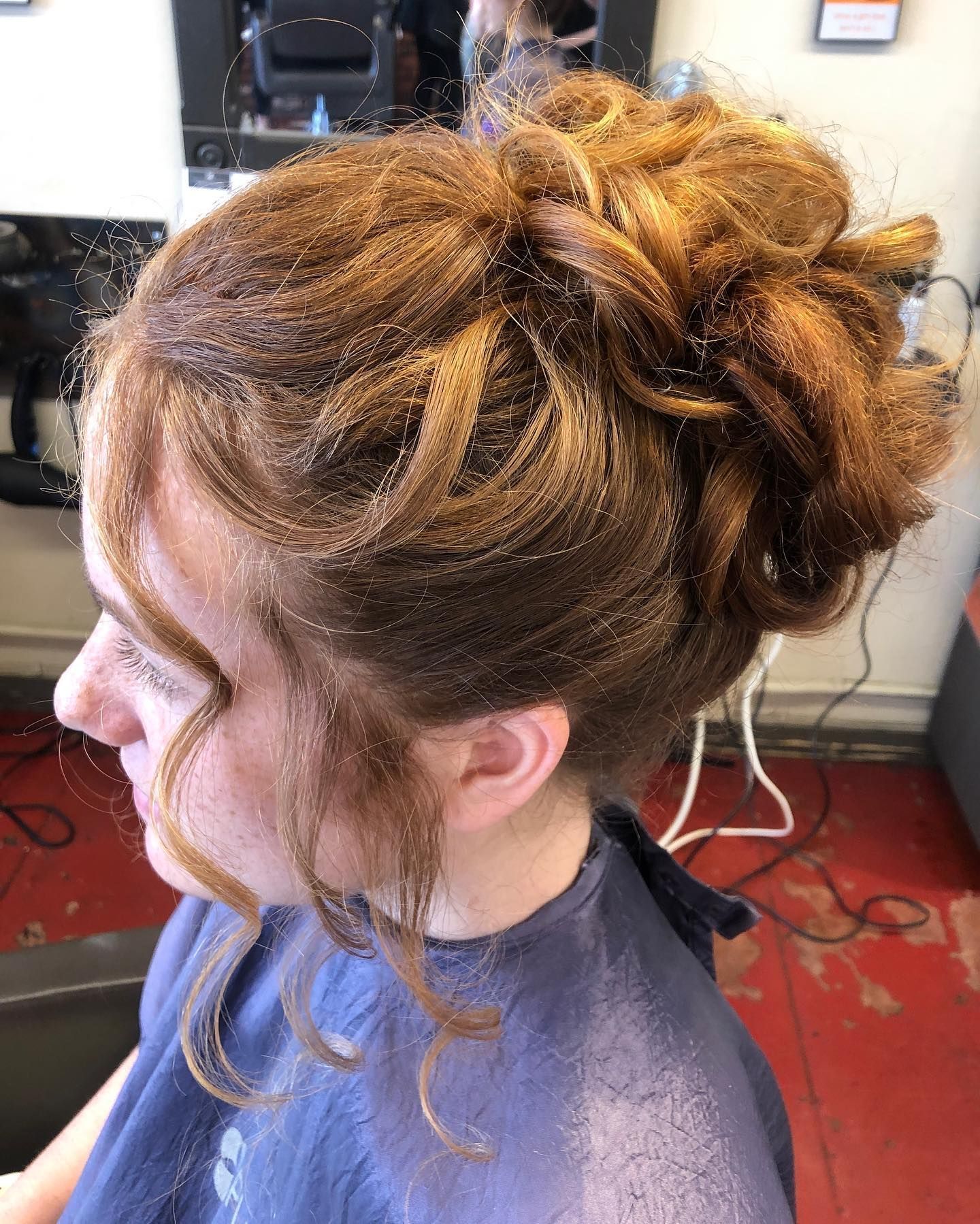 Woman with auburn hair in an updo hairstyle. Side profile with loose curls framing the face, in a salon.