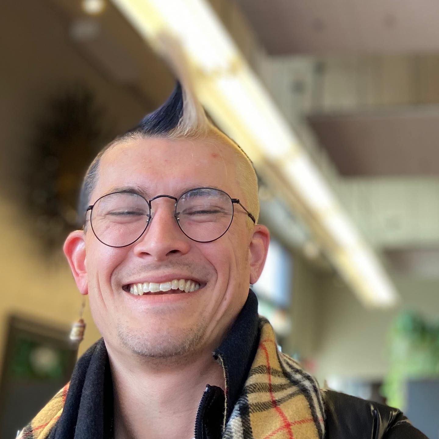 Smiling person with a blue and blonde mohawk, wearing glasses, a scarf, and a leather jacket. Blurred indoor background.