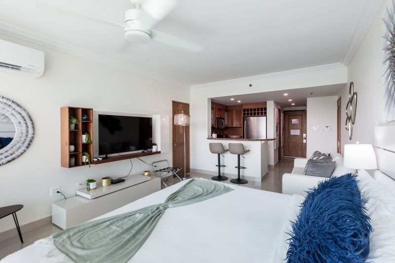 Bright modern bedroom with white bedding, wall-mounted TV, and a view into a kitchen area