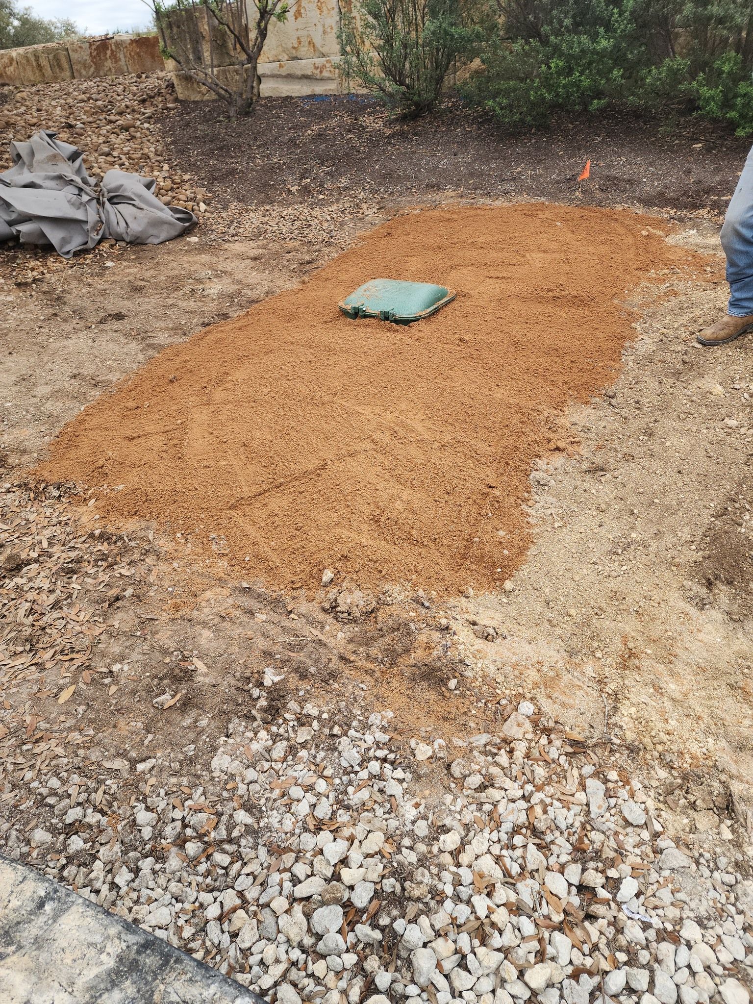A rectangular mulch bed, with a green box, set between gravel and dirt.