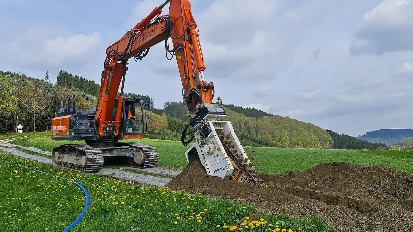 Orange excavator digging a trench in a field near a road on a cloudy day.