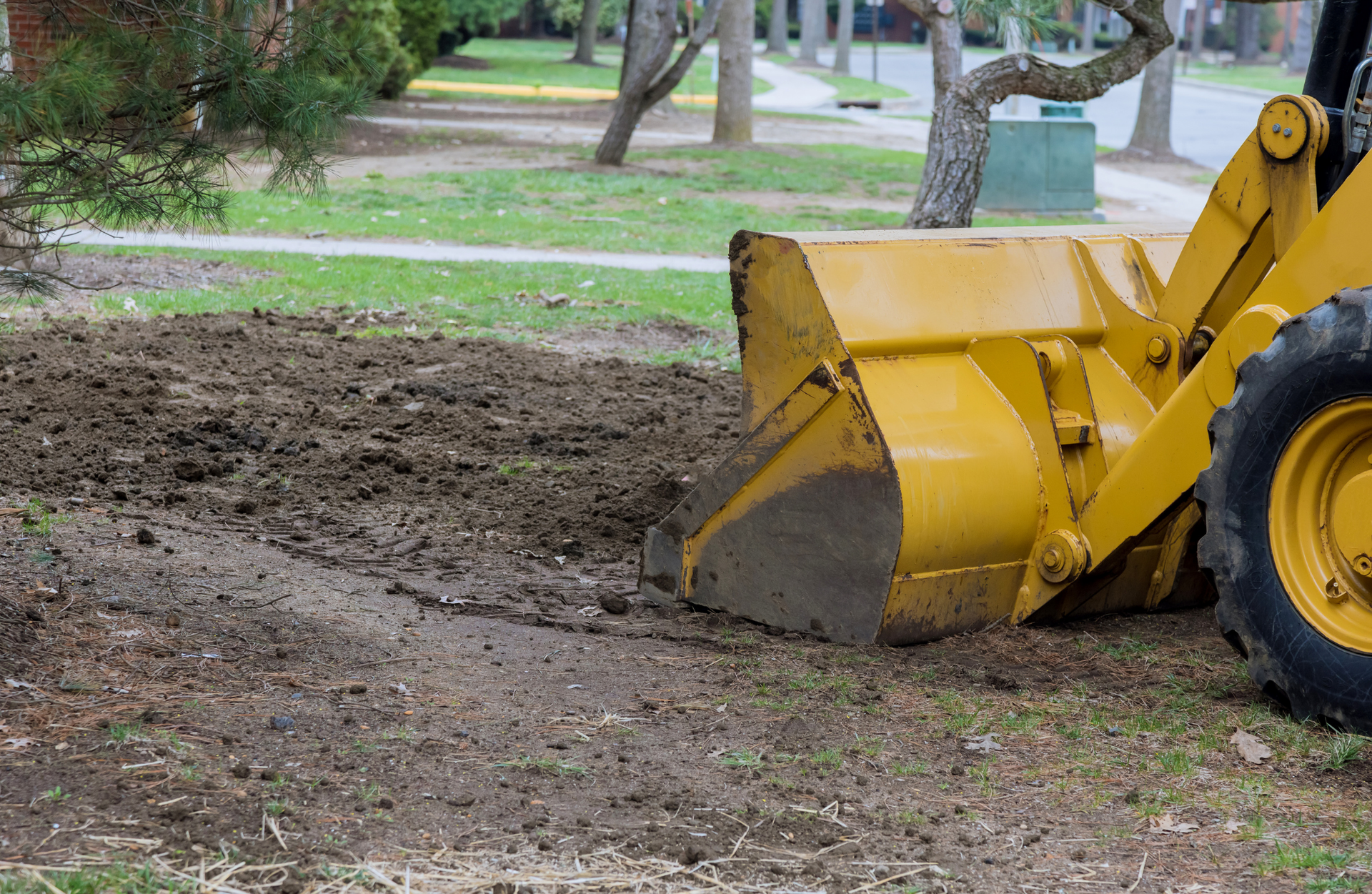 Yellow front-end loader digging into dirt on a residential lawn, preparing for landscaping.