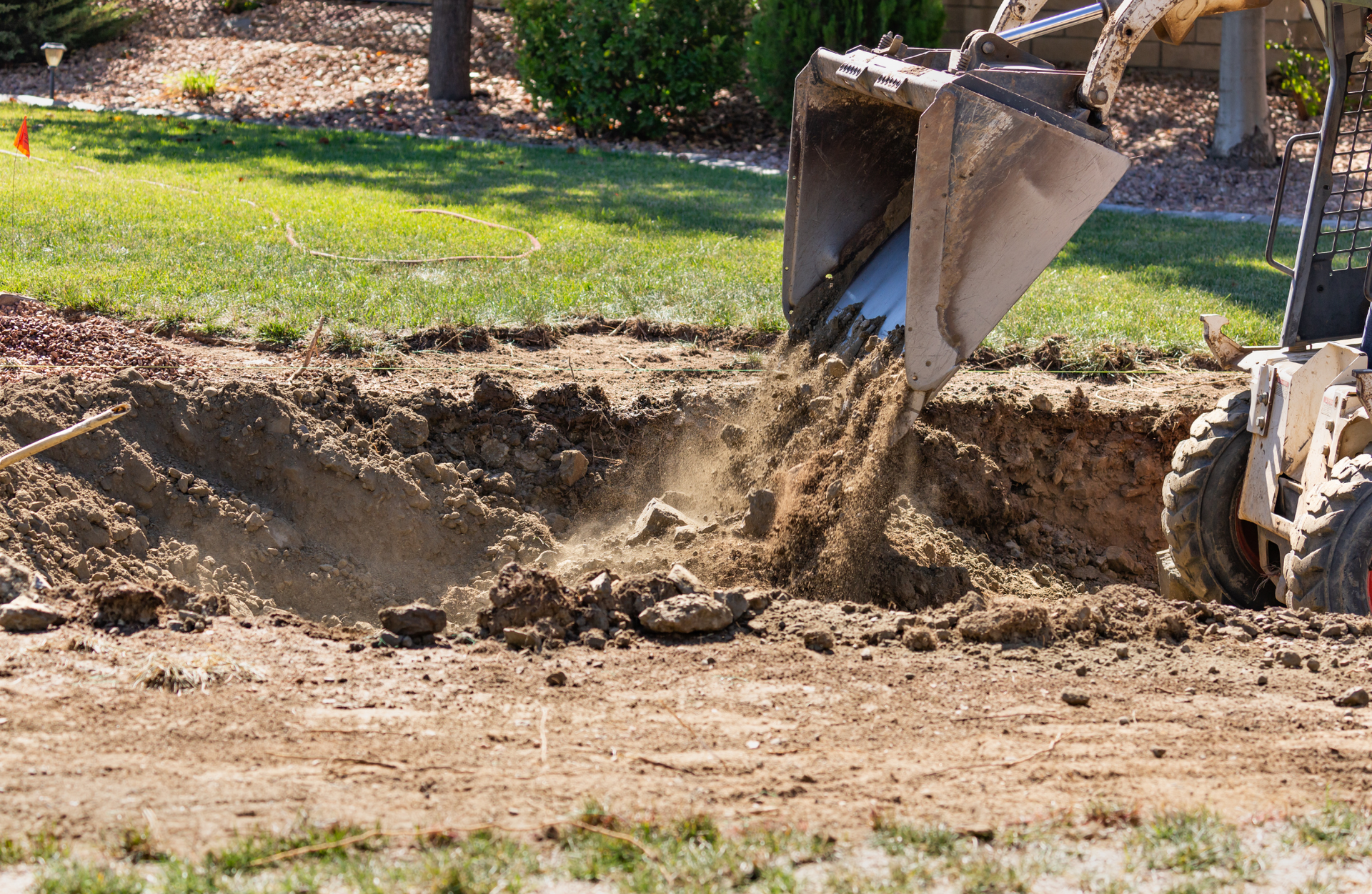 Bobcat excavator dumping dirt from its bucket into a trench in a yard.