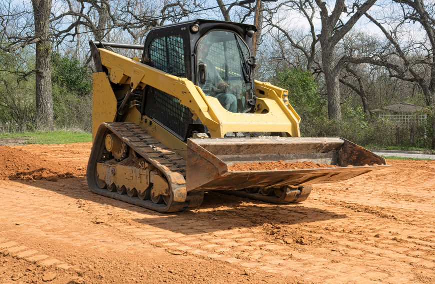 Yellow track loader on reddish-brown dirt, clearing a flat area near trees.