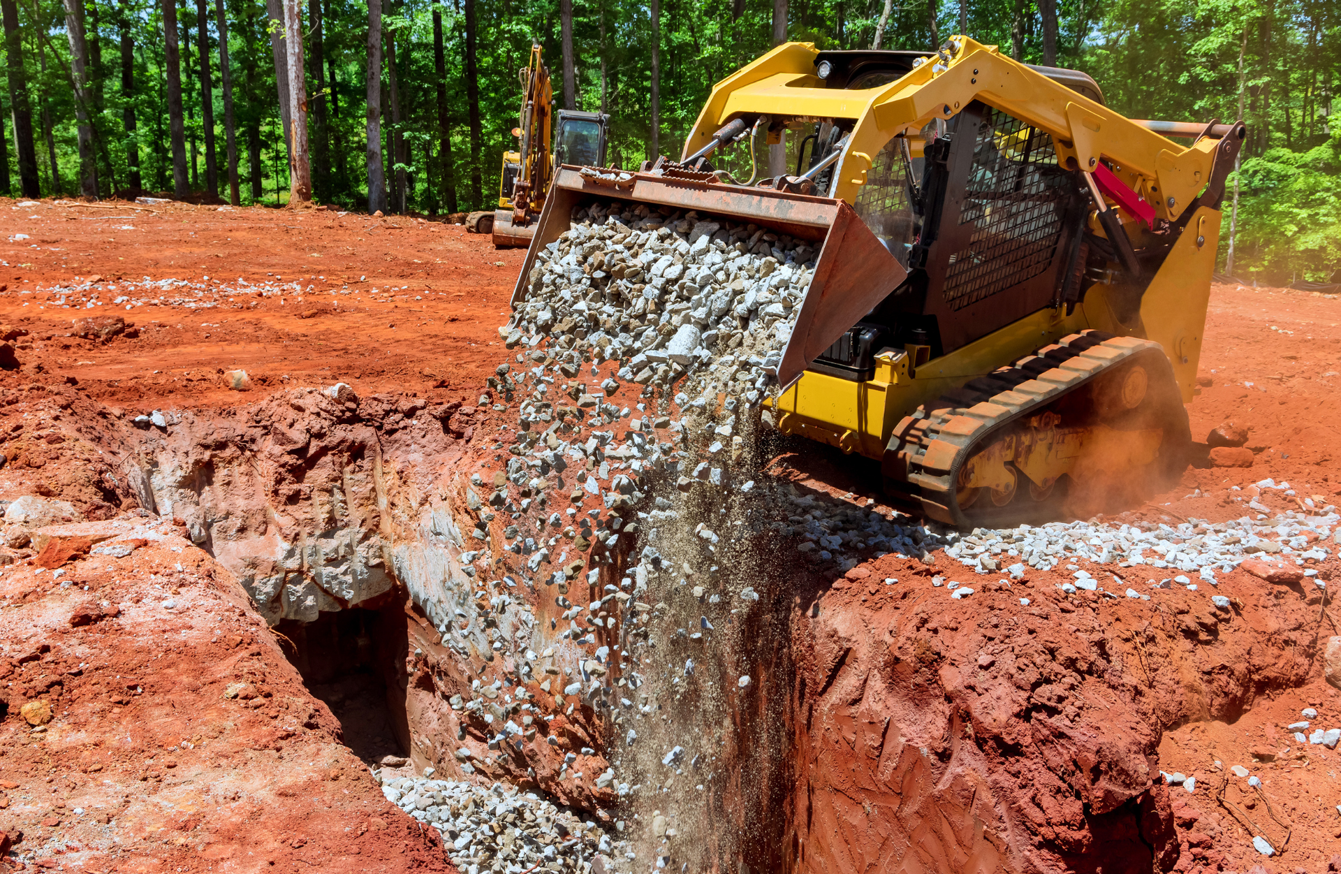 Yellow skid steer dumping gravel into a trench at a construction site.