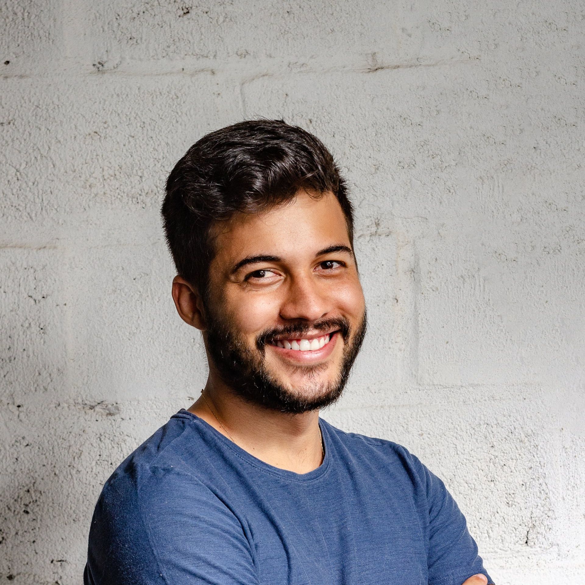 A man with a beard wearing a blue shirt is smiling with his arms crossed.