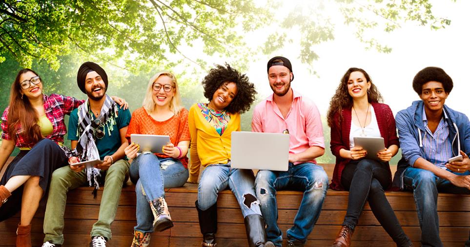 Group of people sitting outdoors, smiling at camera; some with devices. Sunny, tree background.