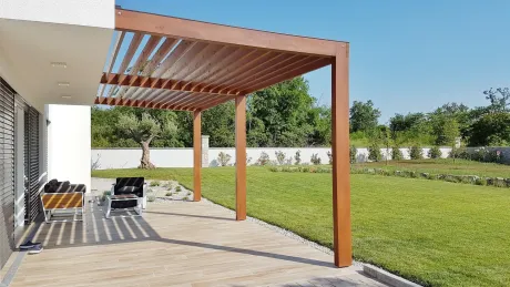 Brown pergola over a patio, casting shadows on the stone tiles. A grassy yard and trees are in the background.