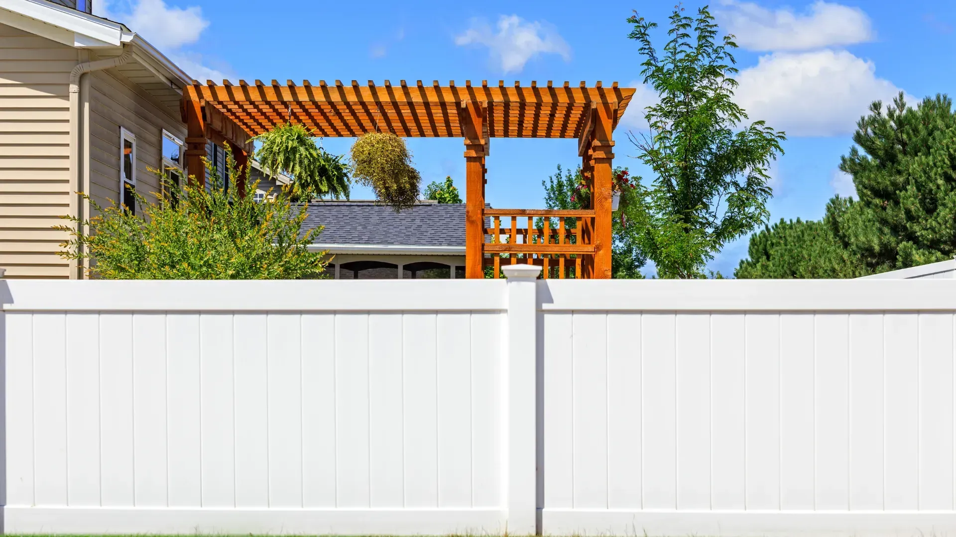White privacy fence in front of a wooden pergola, house, and trees under a blue sky.