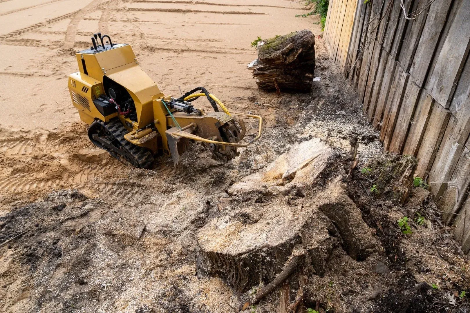 Yellow excavator clearing mud and debris beside a wooden fence after flooding
