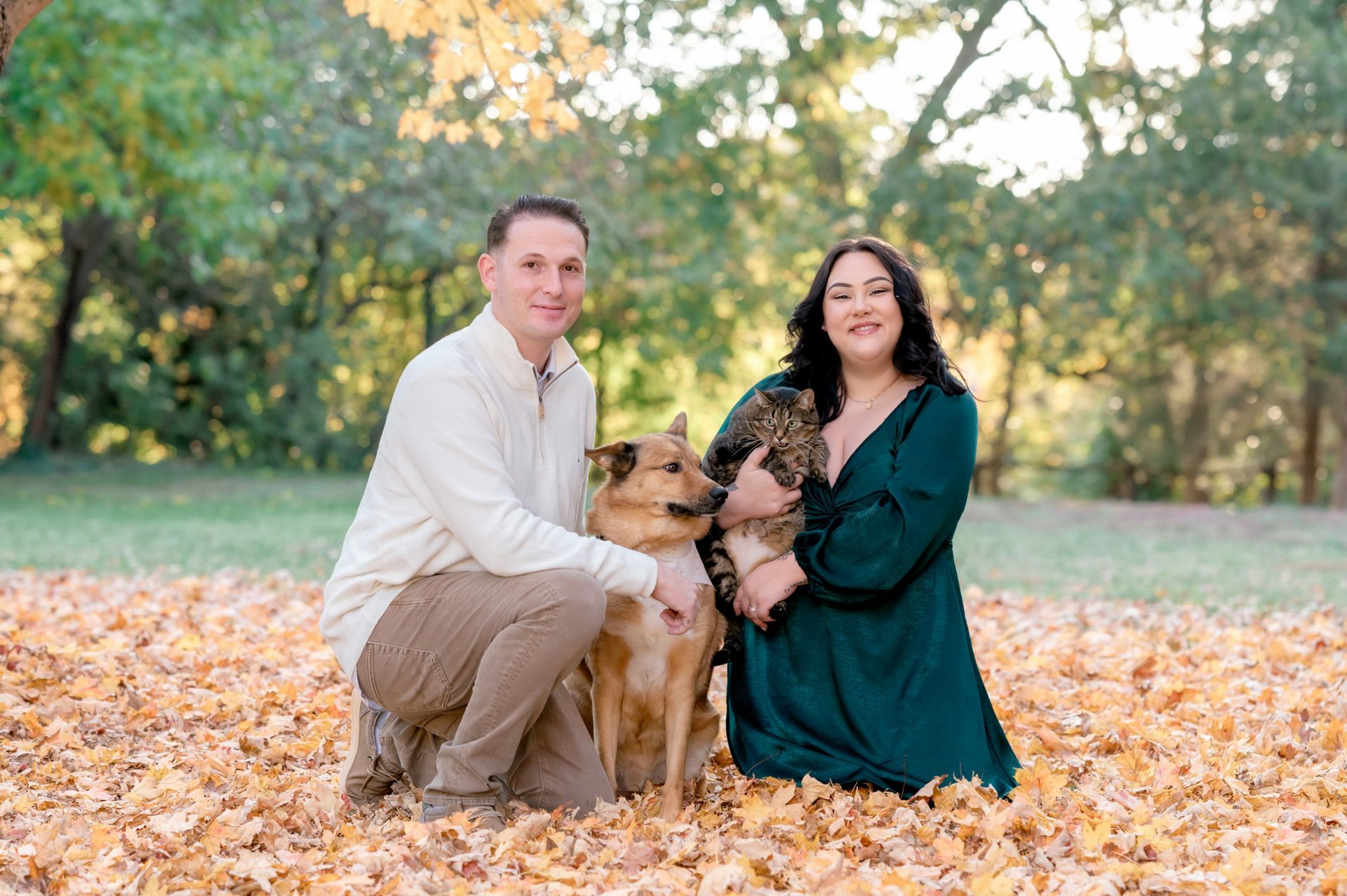 A person and a partner kneel in autumn leaves outdoors with a dog and a cat in their arms, smiling at the camera.