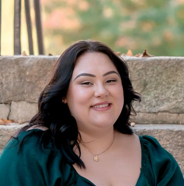 A person with dark hair wearing a green top and a gold necklace, smiling while sitting in front of a stone wall.