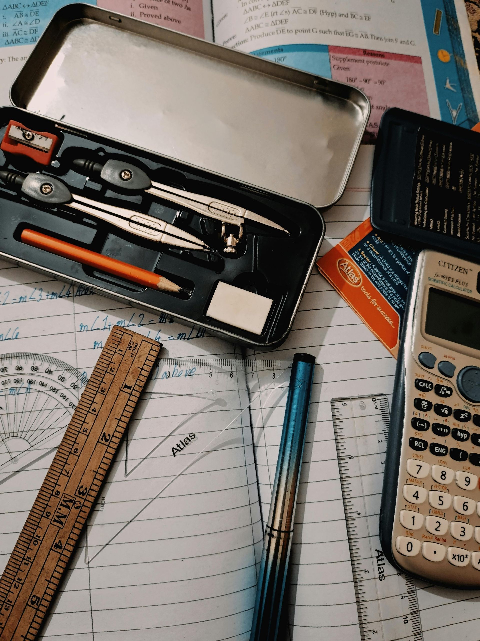 A geometry set, a calculator, a wooden ruler, and a pen arranged on a desk over open notebook pages.