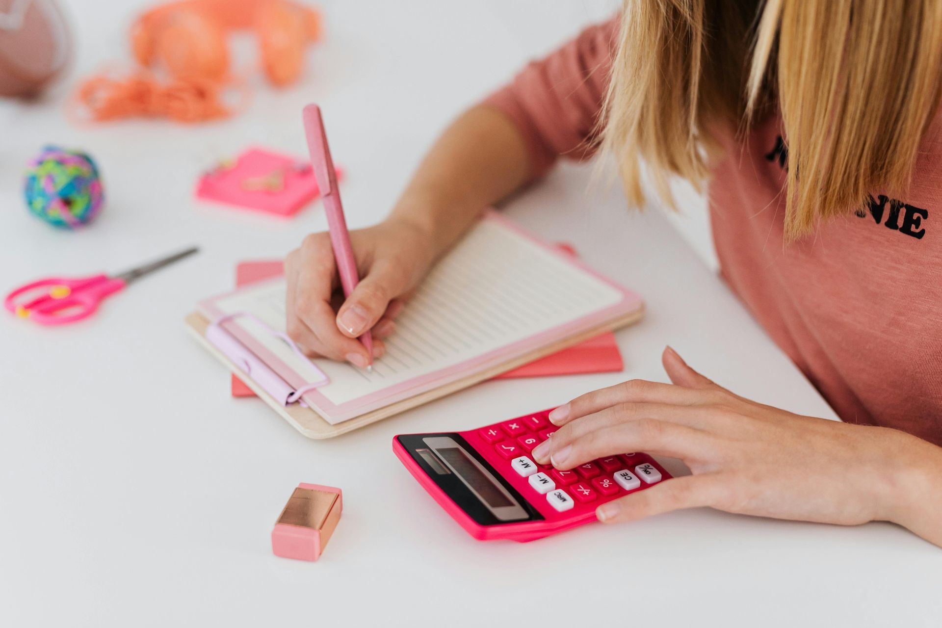 A person at a white desk writing on a notepad with a pink pen while using a pink calculator. Desk supplies are nearby.