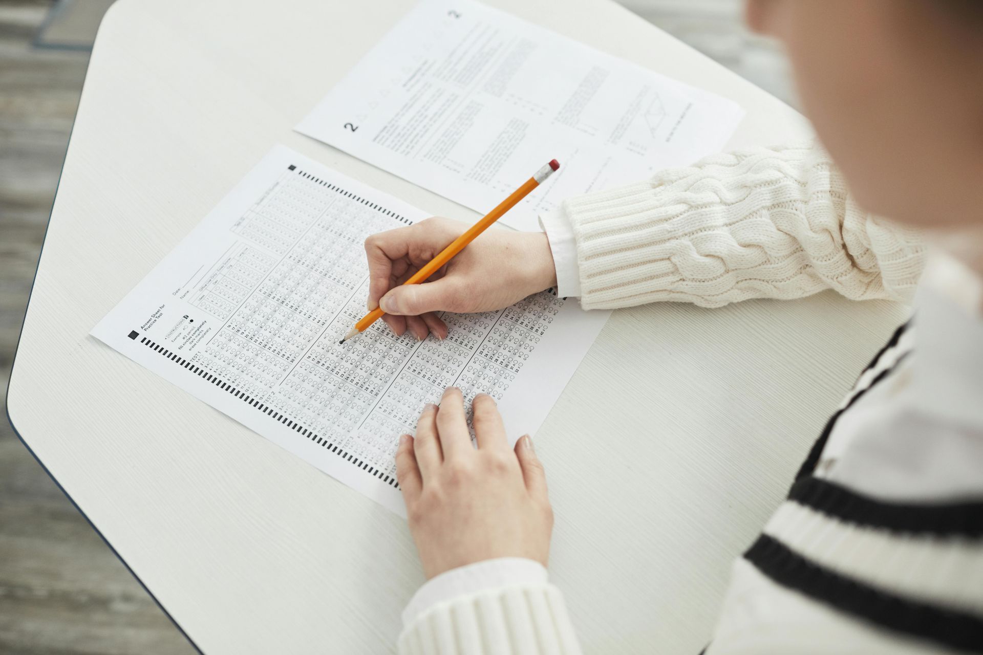 A person wearing a striped sweater fills in a grid-style bubble sheet with a pencil at a white desk.