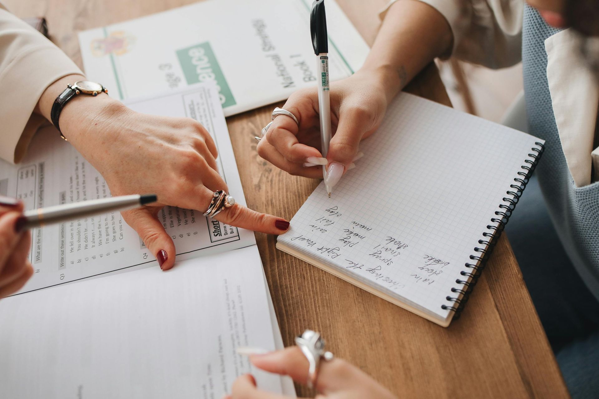 Two people seated at a wooden desk collaborating on work, one writing in a notebook while the other points to a document.