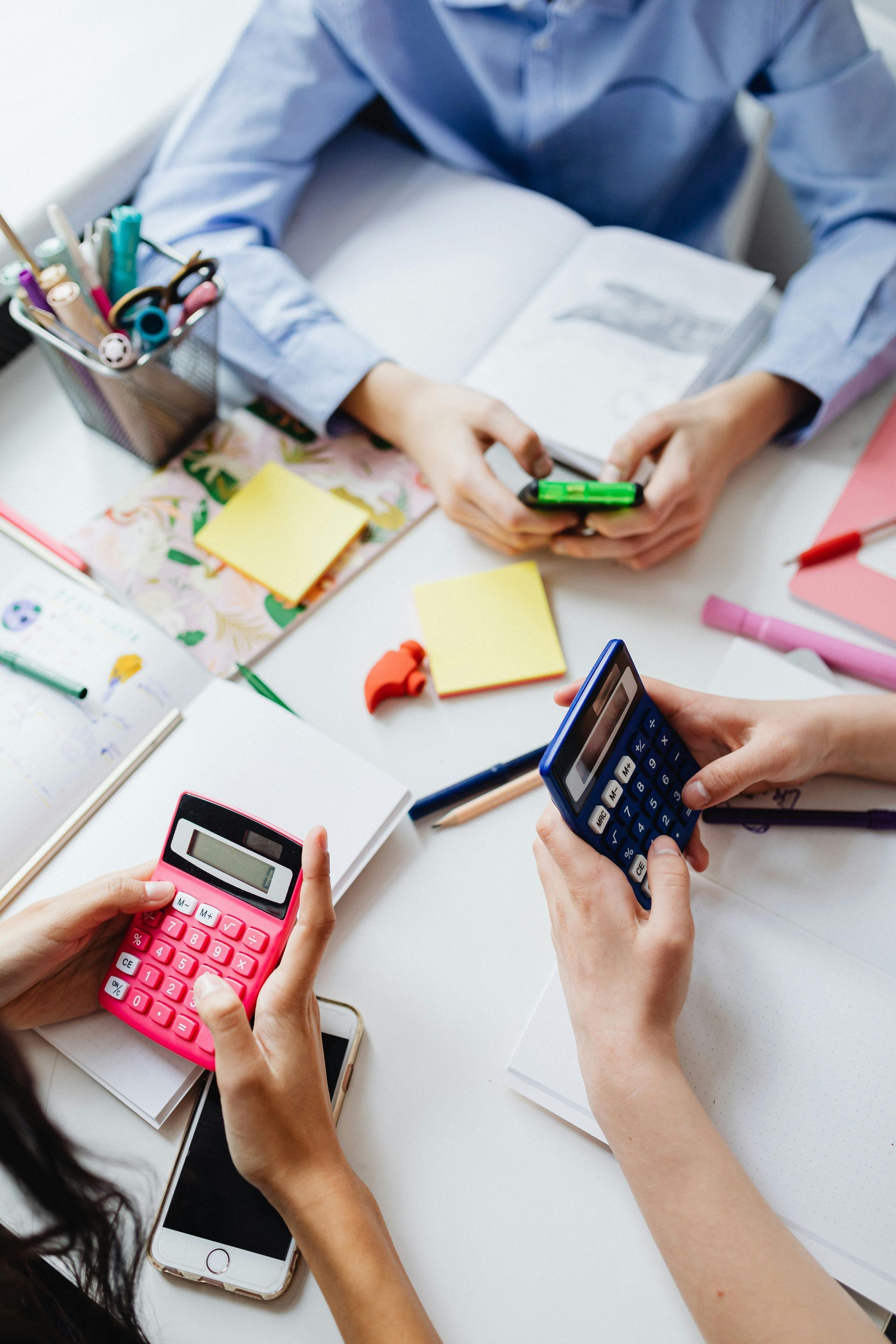 Three people sit around a white table studying with calculators, smartphones, and notebooks.