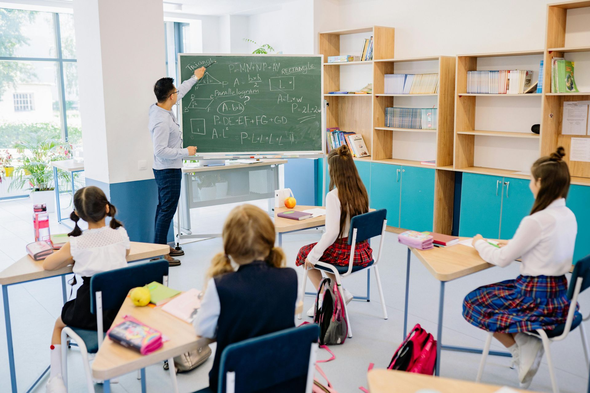 A teacher writes on a blackboard in a classroom while students sit at individual desks facing the board.
