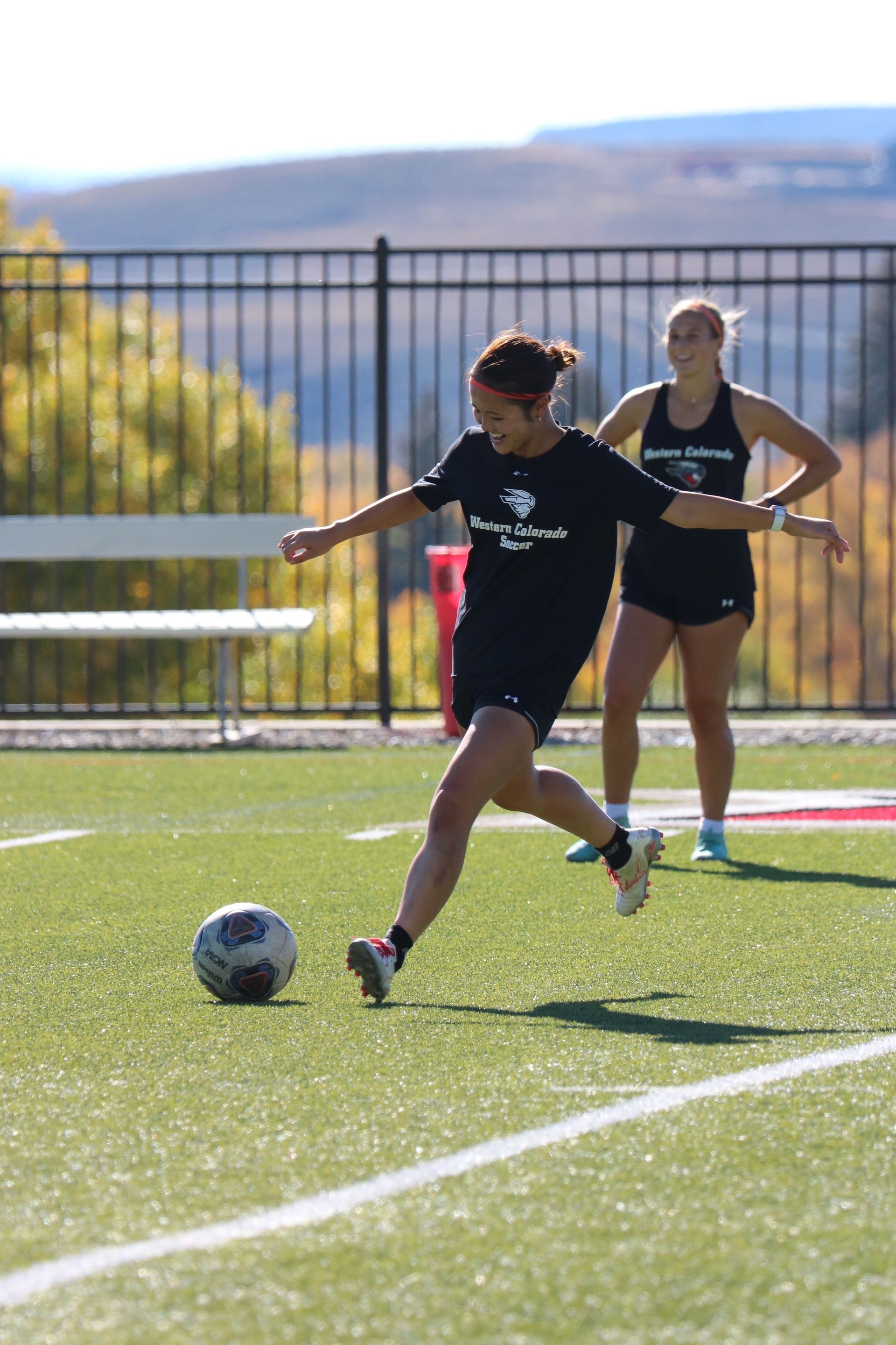 A woman is kicking a soccer ball on a field while another woman watches.