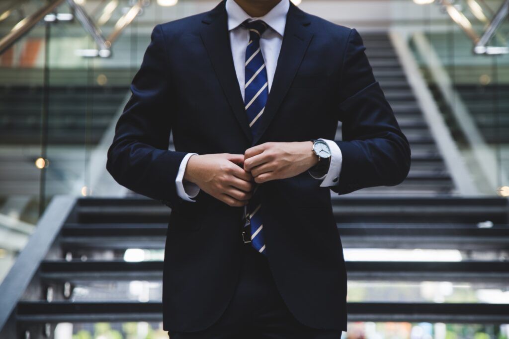 Man in navy suit buttoning jacket, striped tie, standing near staircase.