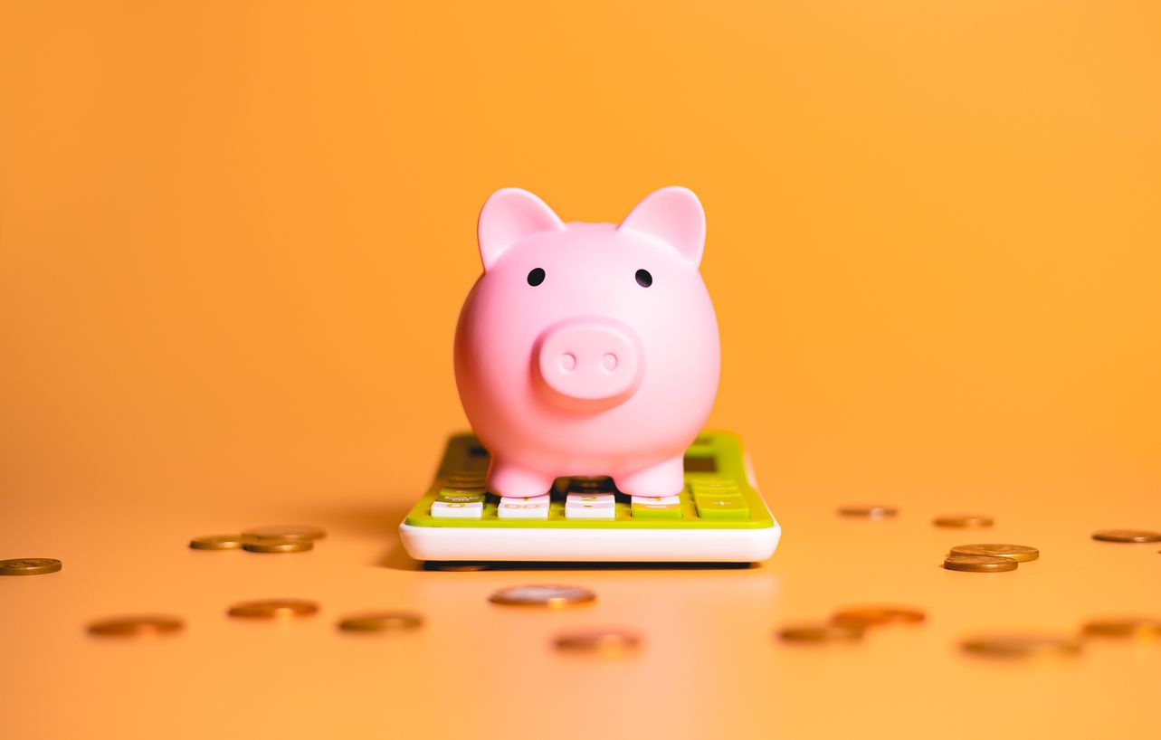 Pink piggy bank on a calculator, surrounded by scattered coins, against a yellow background.
