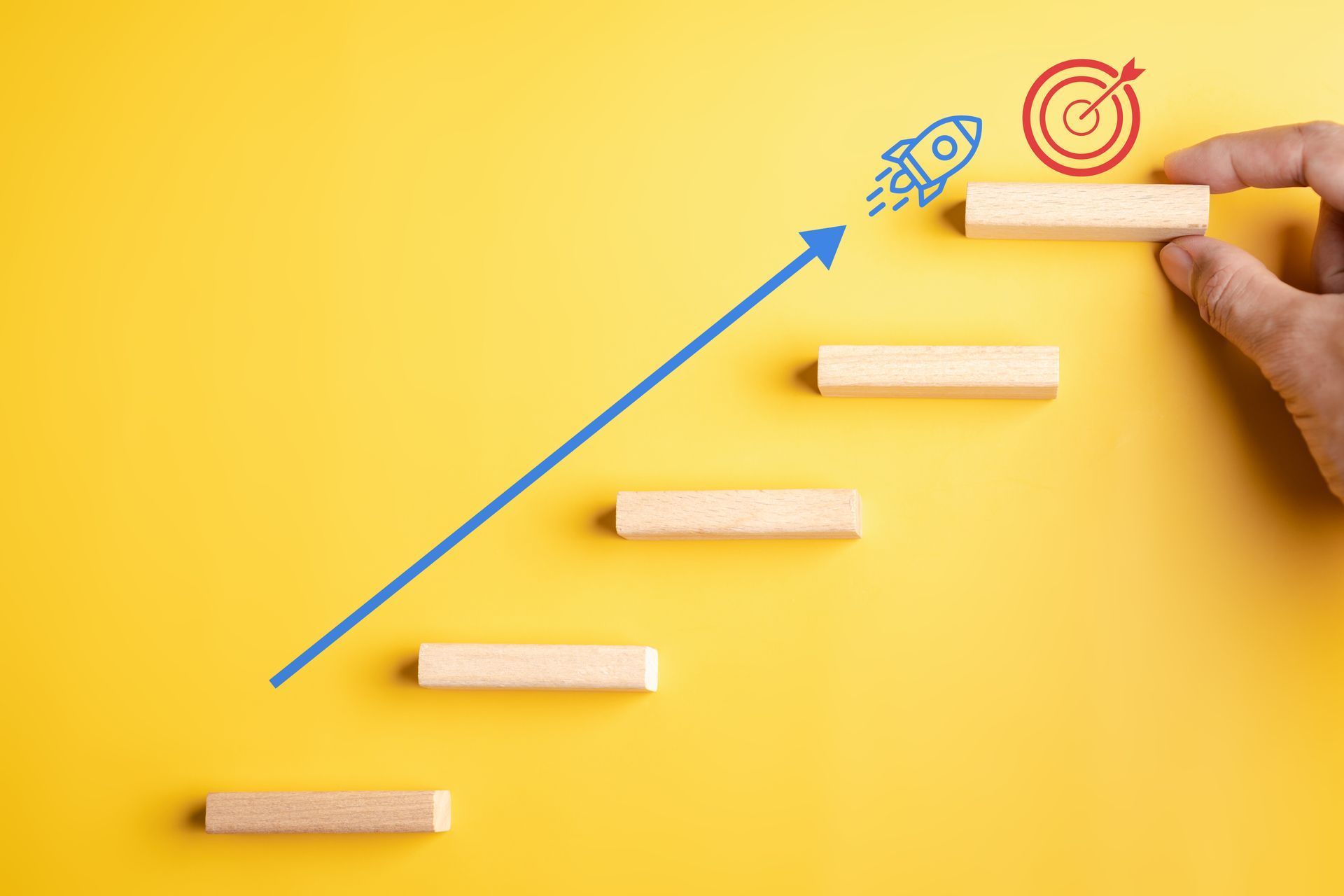 Hand placing wooden block on staircase with upward arrow, rocket, and target.