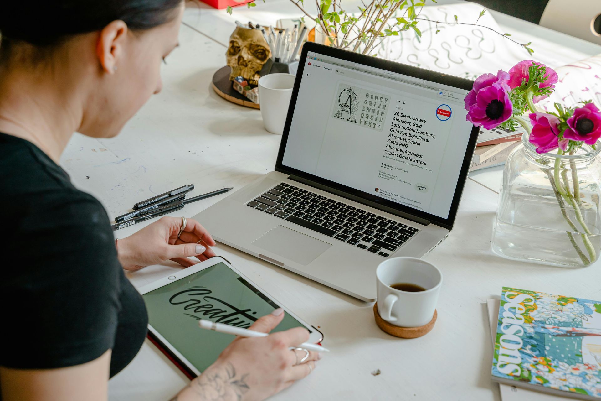 Woman working on laptop and tablet at a white table, with flowers and coffee.