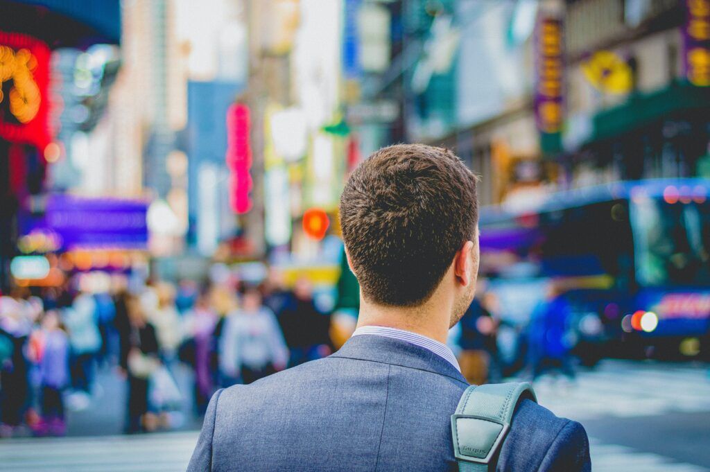 Man in a suit looks toward a crowded street scene with bright signs and buildings.