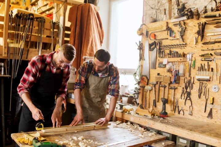 Two people in a workshop measuring wood with tools, surrounded by equipment.