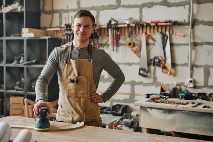 Man in apron, sanding wood in workshop, smiling. Tools and worktable in the background.