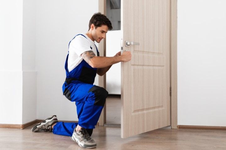 Person kneeling, installing a door handle in a room. Wearing blue overalls, white shirt.