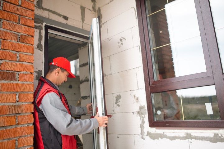 Construction worker installing a door frame into a brick and concrete wall beside a window.
