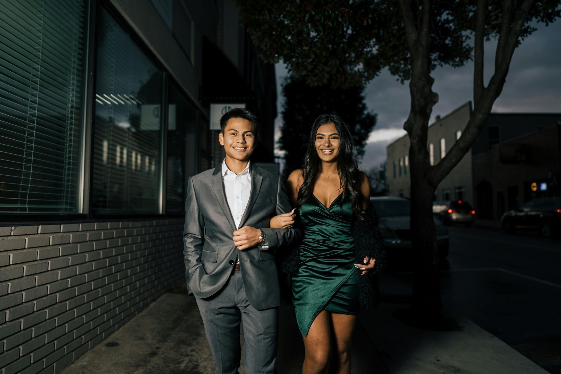 Couple walking arm in arm, smiles, at night. Man in gray suit, woman in green dress. Building and street in background.