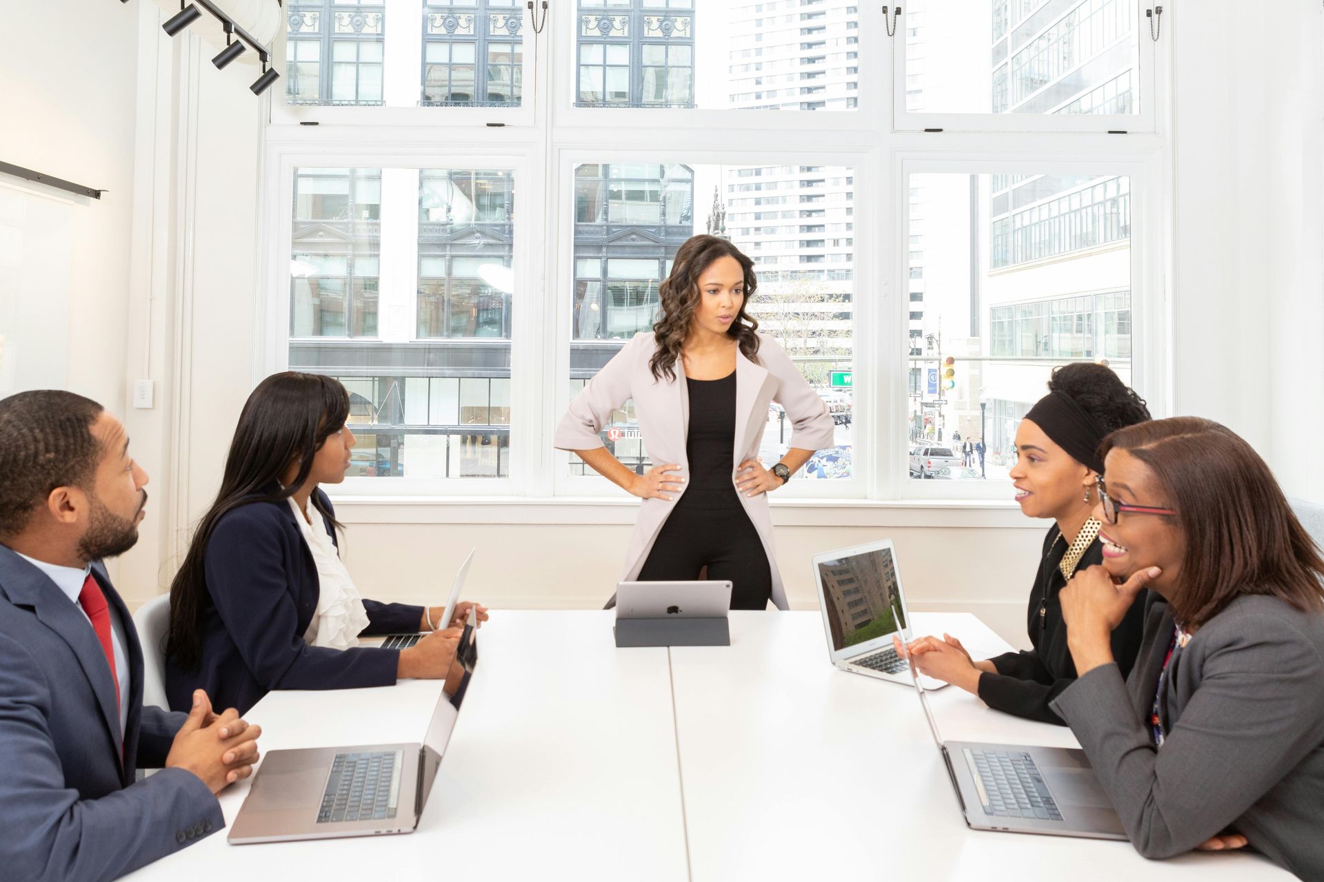 Business team meeting in a bright office; woman stands, arms akimbo, addressing the group at a long table.