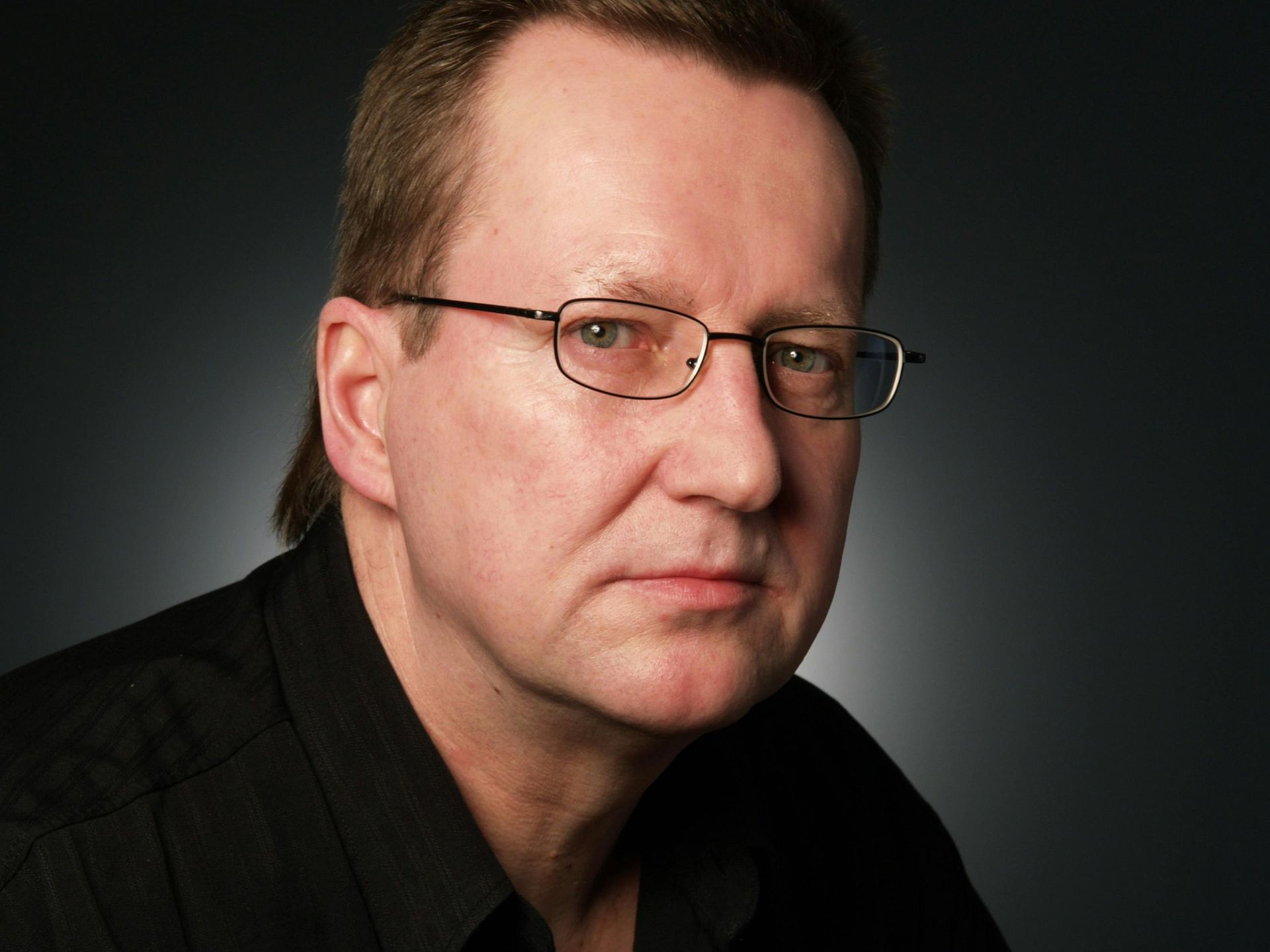 Man with glasses, wearing a black shirt, looking slightly to the side. Dark background.