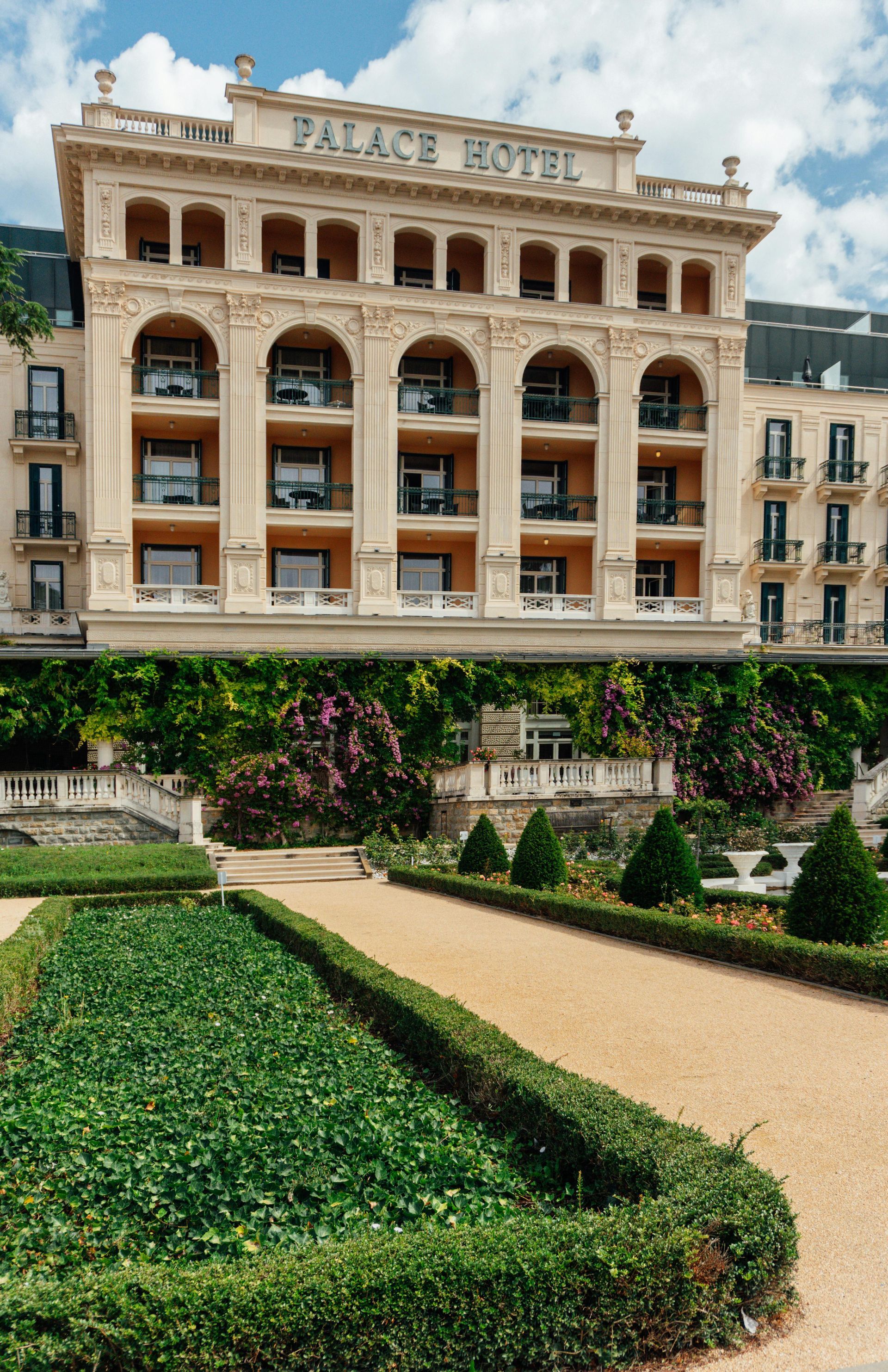 Palace Hotel facade, ornate architecture above manicured garden, tan pathway, green hedges.