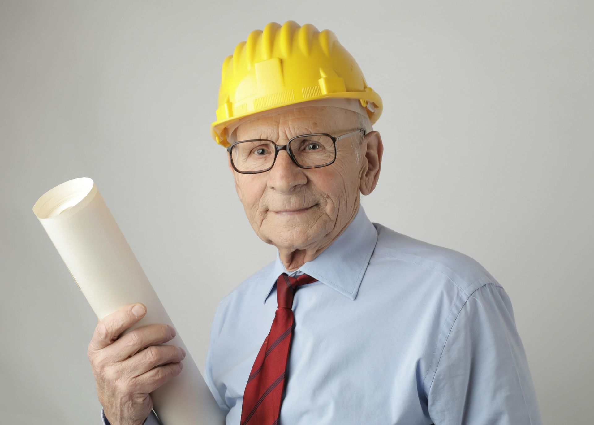 Elderly man in a yellow hard hat and glasses, holding rolled blueprints, wearing a red tie and blue shirt.
