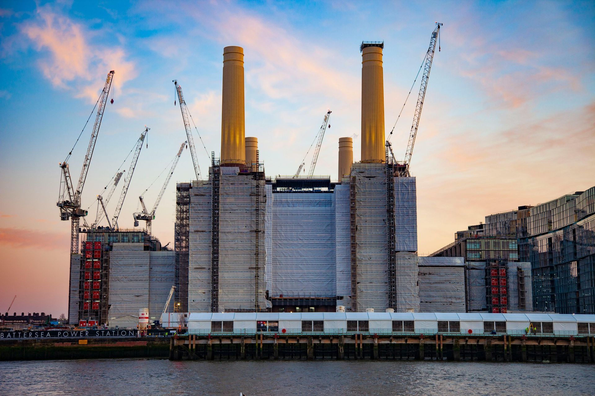 Battersea Power Station under construction, riverside. Grey facade, four yellow chimneys, cranes, and cloudy sky.