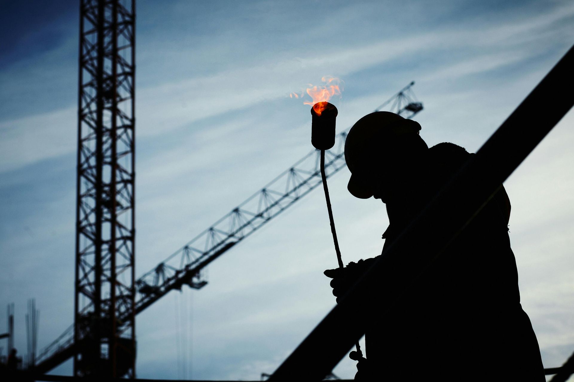 Silhouette of construction worker welding on steel beam, crane in background.