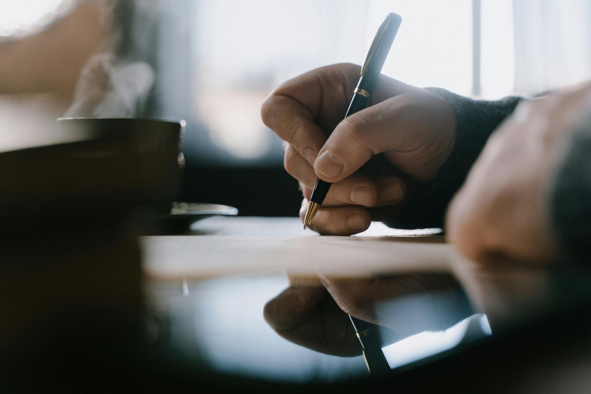 Close-up of a hand writing with a pen on paper; a coffee cup is next to the hand, indoors.