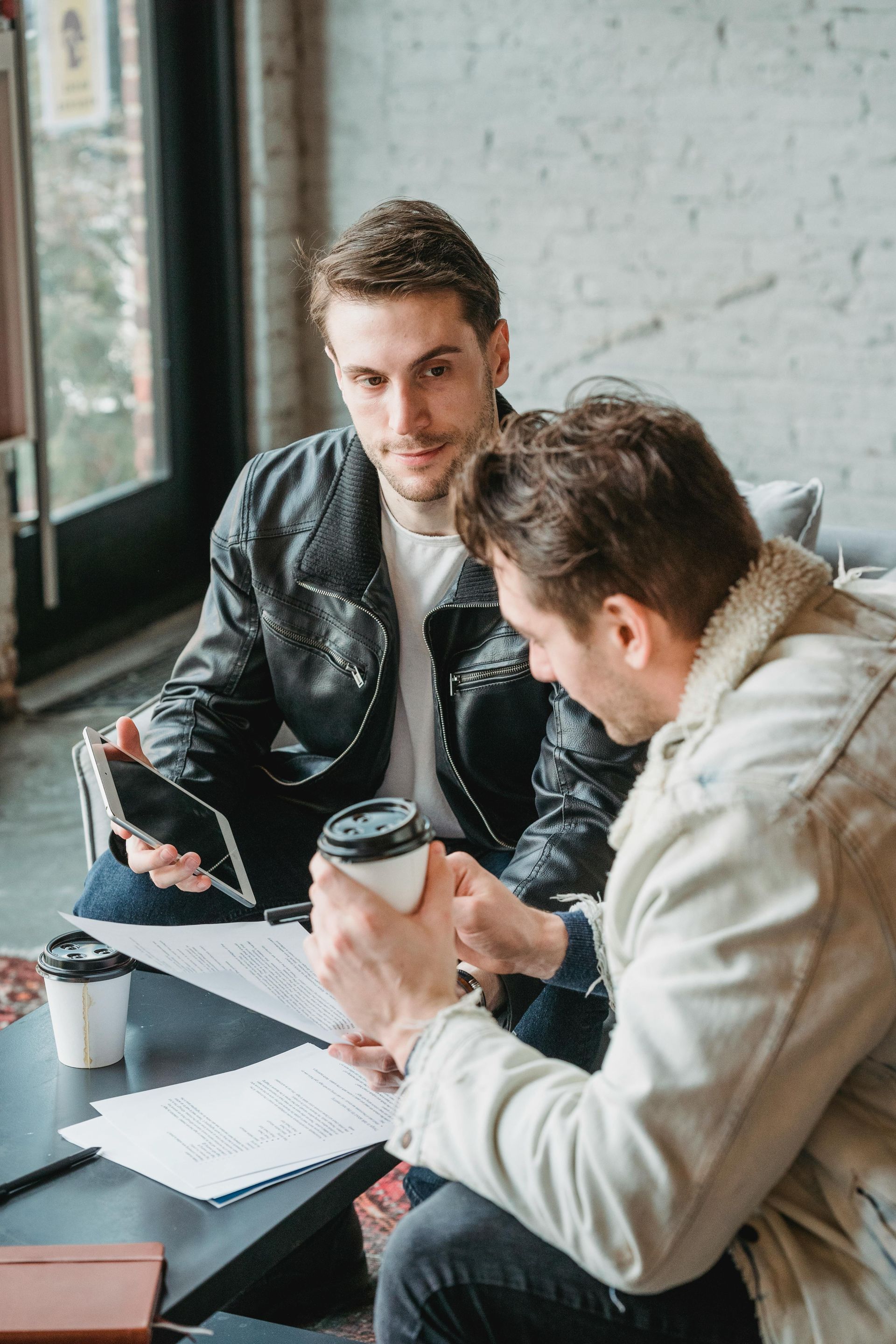 Two men at table, reviewing papers, one holding a tablet and another a coffee cup.