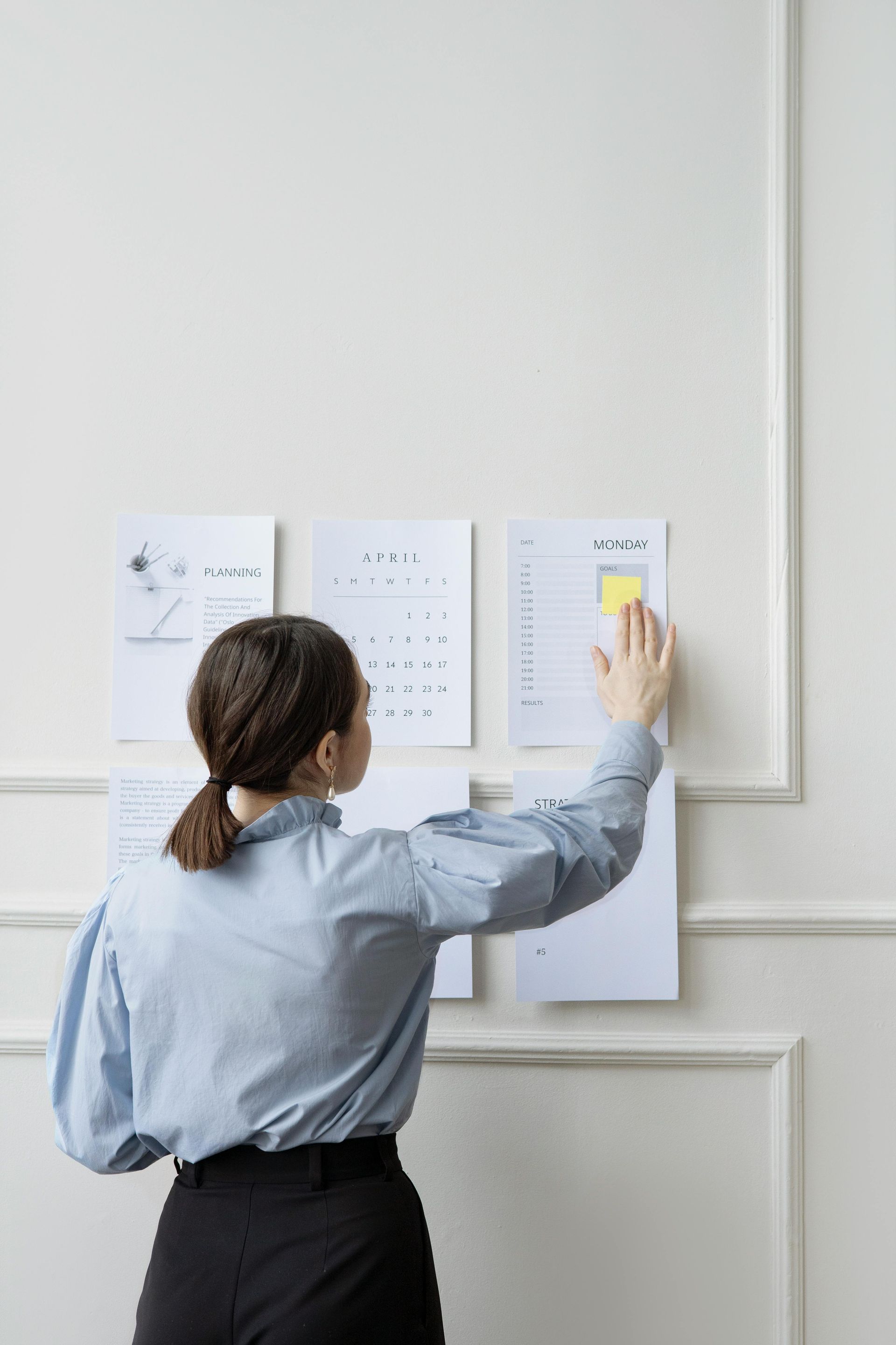 Woman in a blue shirt and black pants, putting a sticky note on a wall with papers.