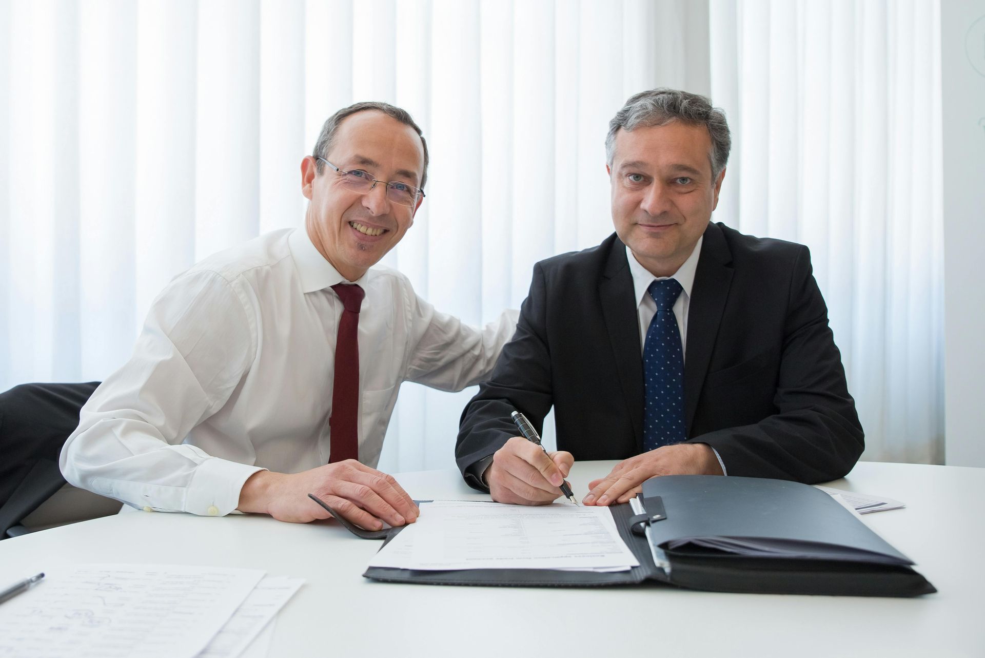 Two men in suits sign a document at a table, smiling. One man has his arm on the other's shoulder.