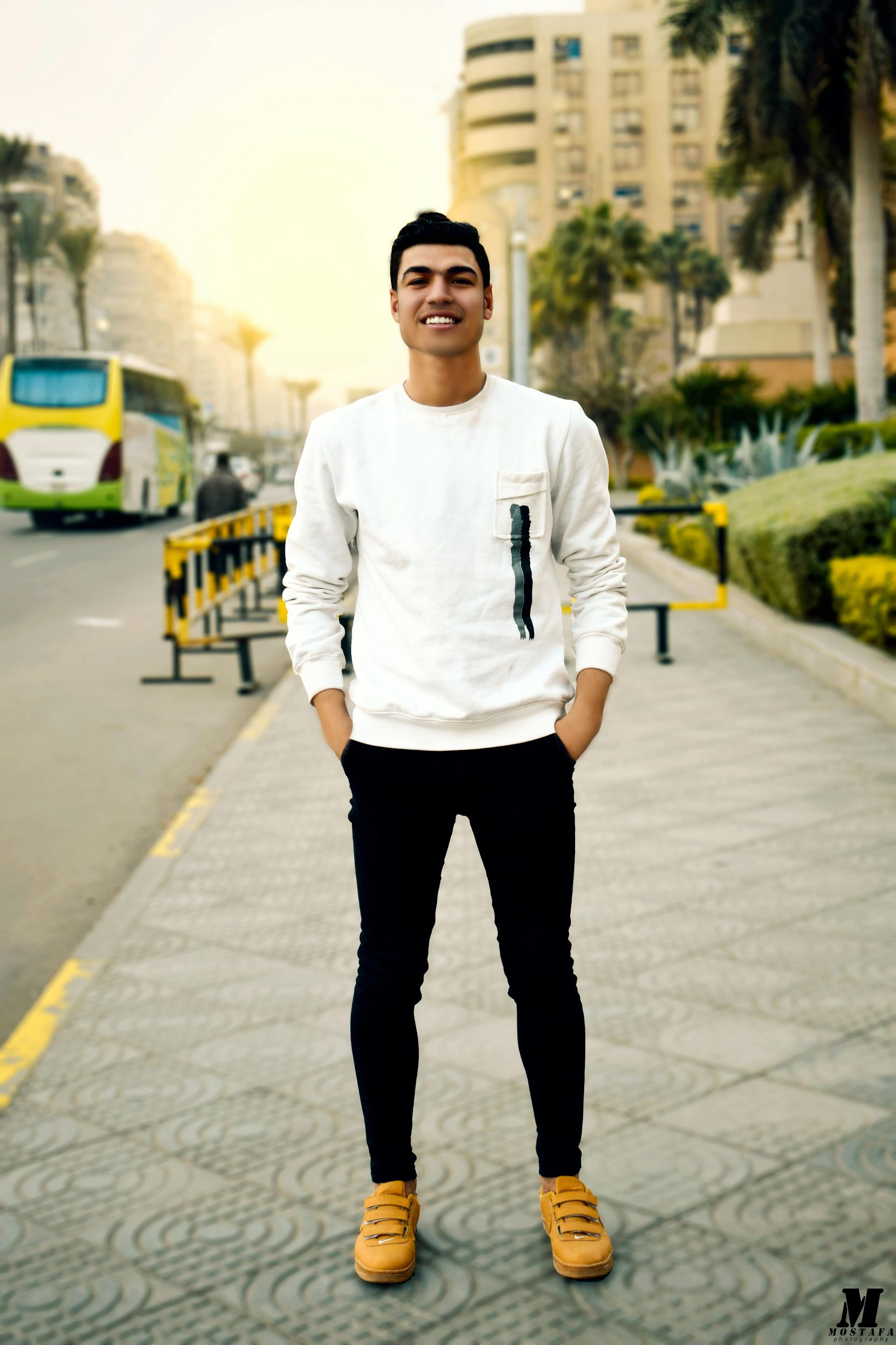 Man standing on a sidewalk, smiling. He wears a white sweater, black pants, and tan shoes. City street background.