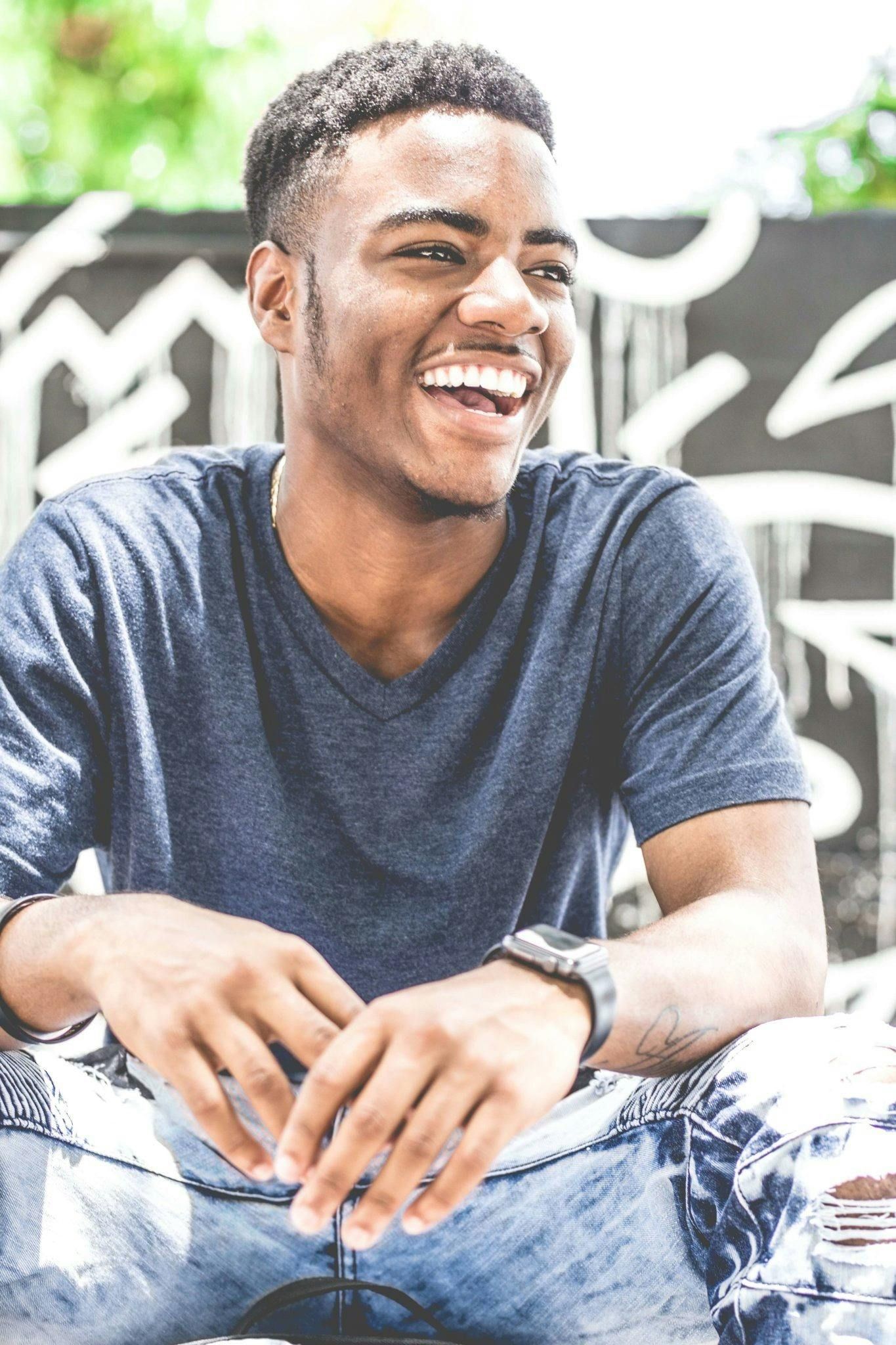 Smiling young Black man, outdoors, wearing blue shirt, watch, leaning forward. Graffiti background.