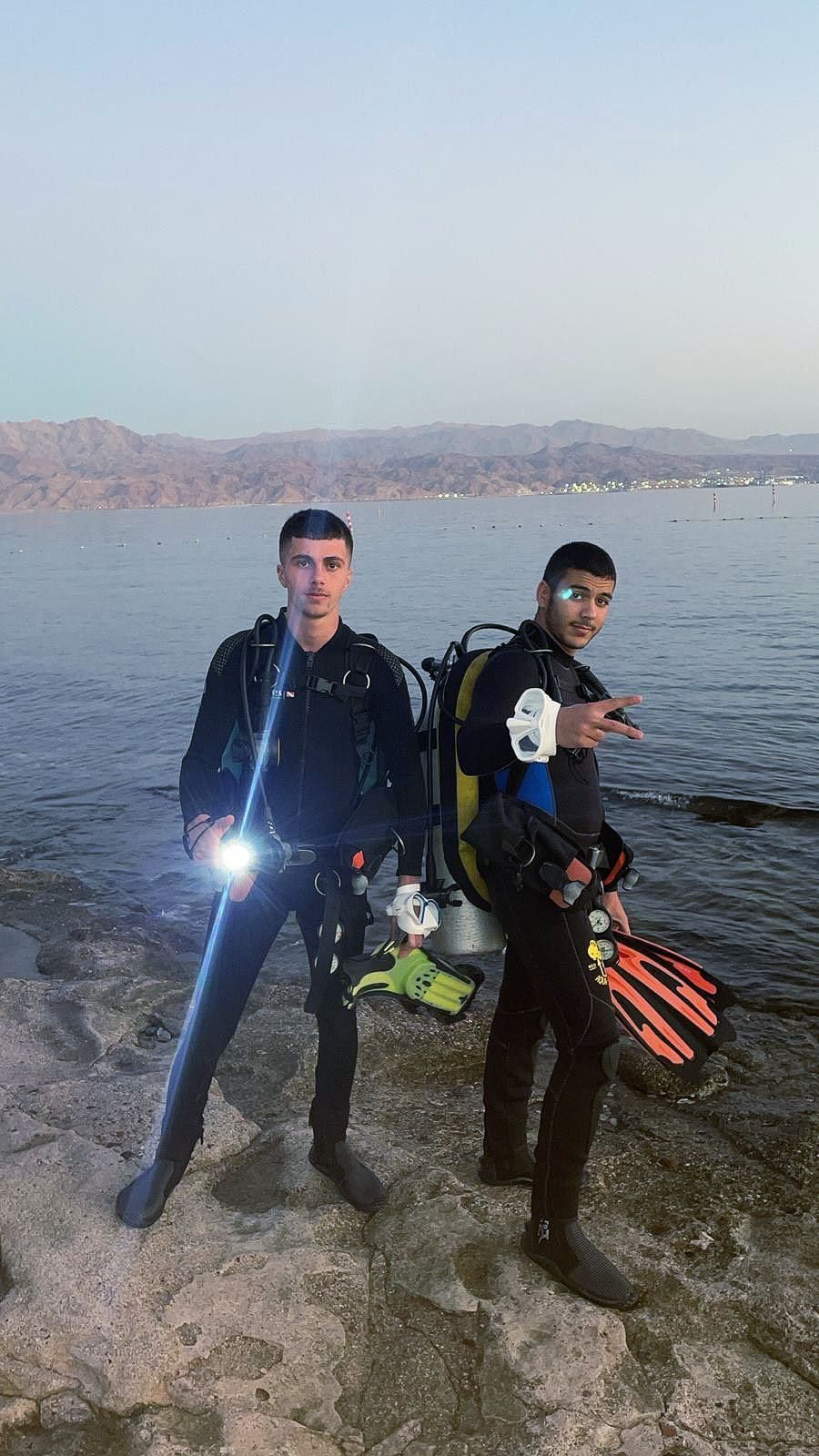 Two scuba divers are standing next to each other on a rock near the water.