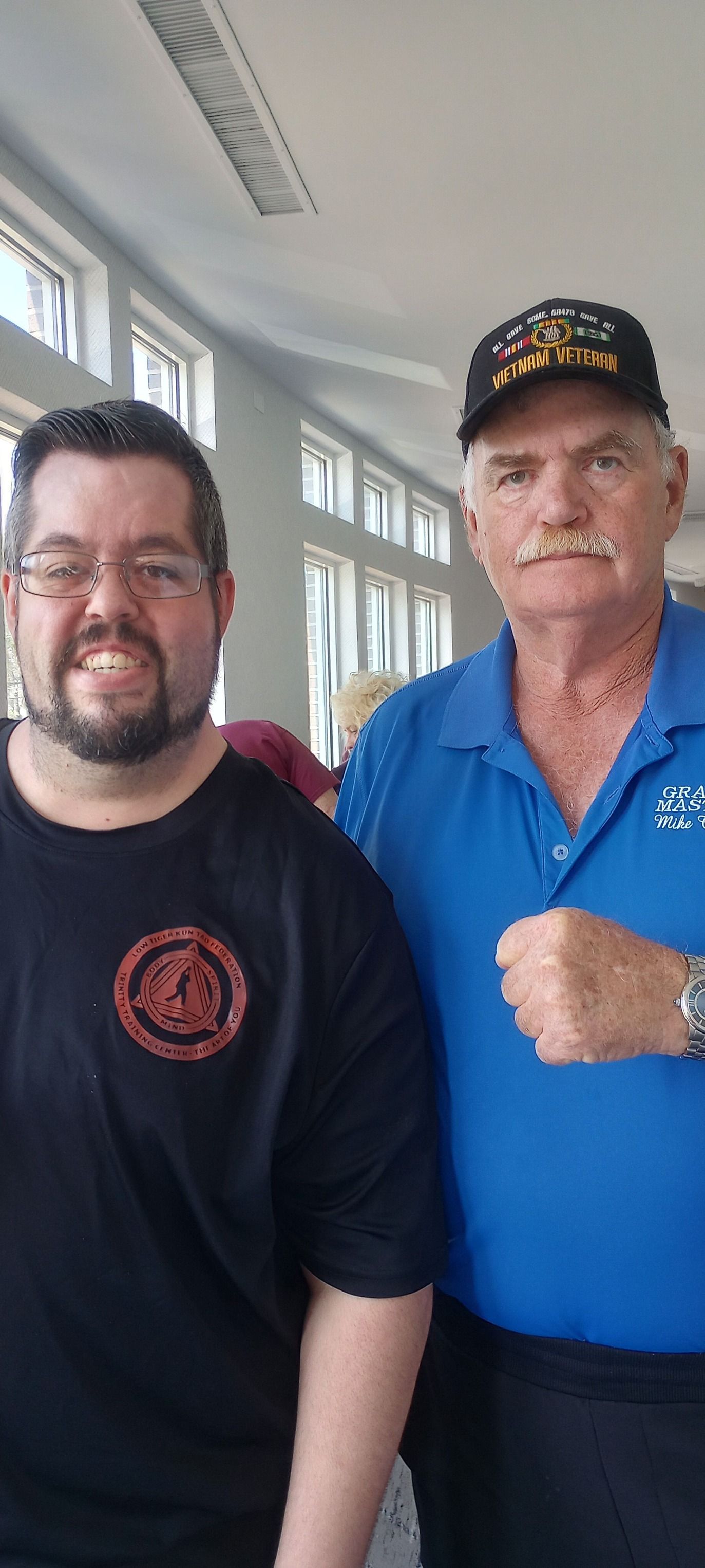 Two men smiling indoors. One wears glasses and a black shirt. The other wears a hat and blue polo.