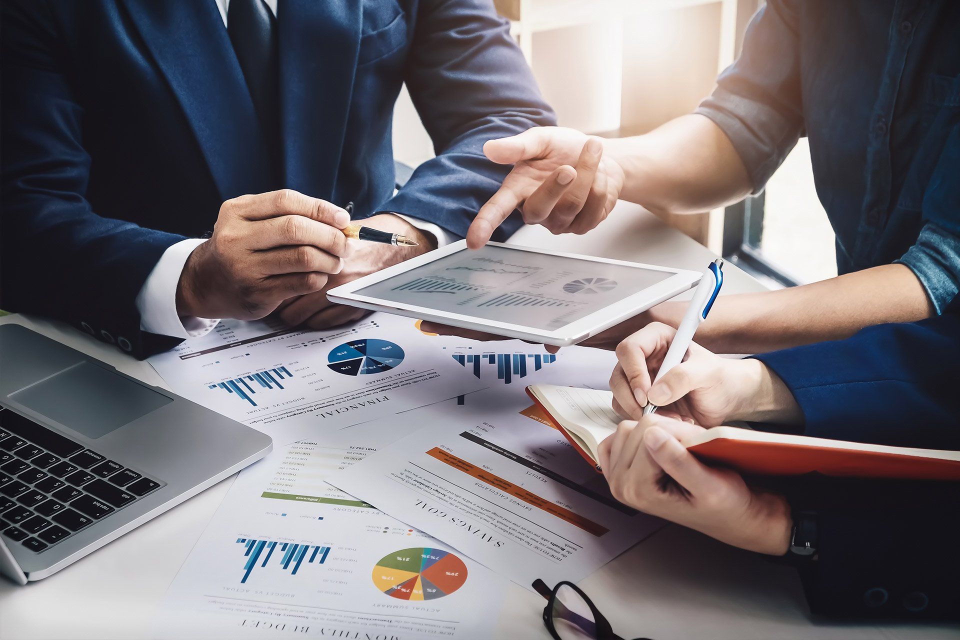 Personas de negocios en una mesa, revisando informes y usando una tableta y una computadora portátil, discutiendo datos.