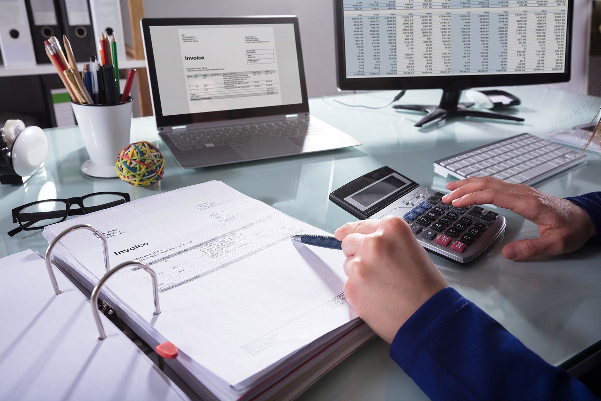 Persona trabajando en un escritorio, usando una calculadora y revisando documentos, con una computadora portátil y un monitor que muestra datos financieros.